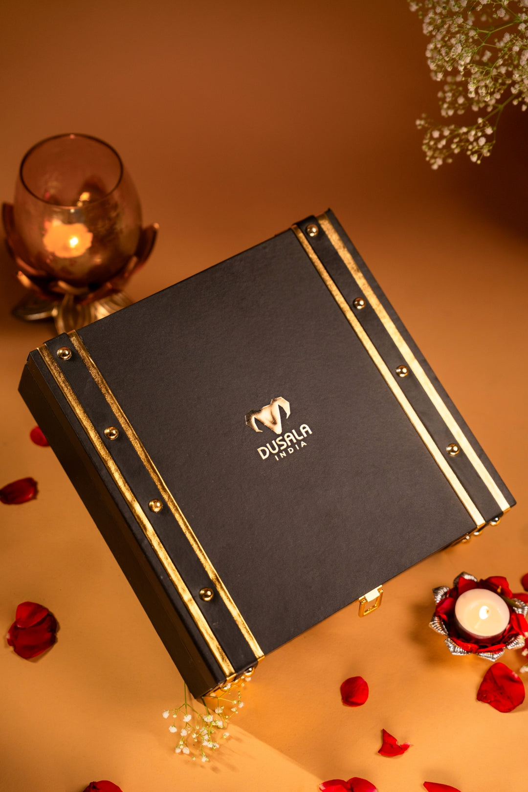 A black rectangular gift box with gold trim and the DUSILA brand logo, styled on a warm beige surface with red rose petals, a lit candle, and soft decorative flowers.