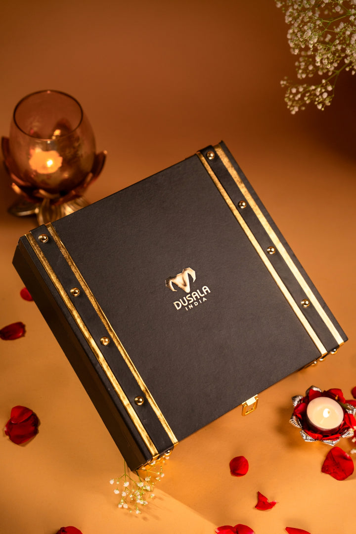 A black rectangular gift box with gold trim and the DUSILA brand logo, styled on a warm beige surface with red rose petals, a lit candle, and soft decorative flowers.